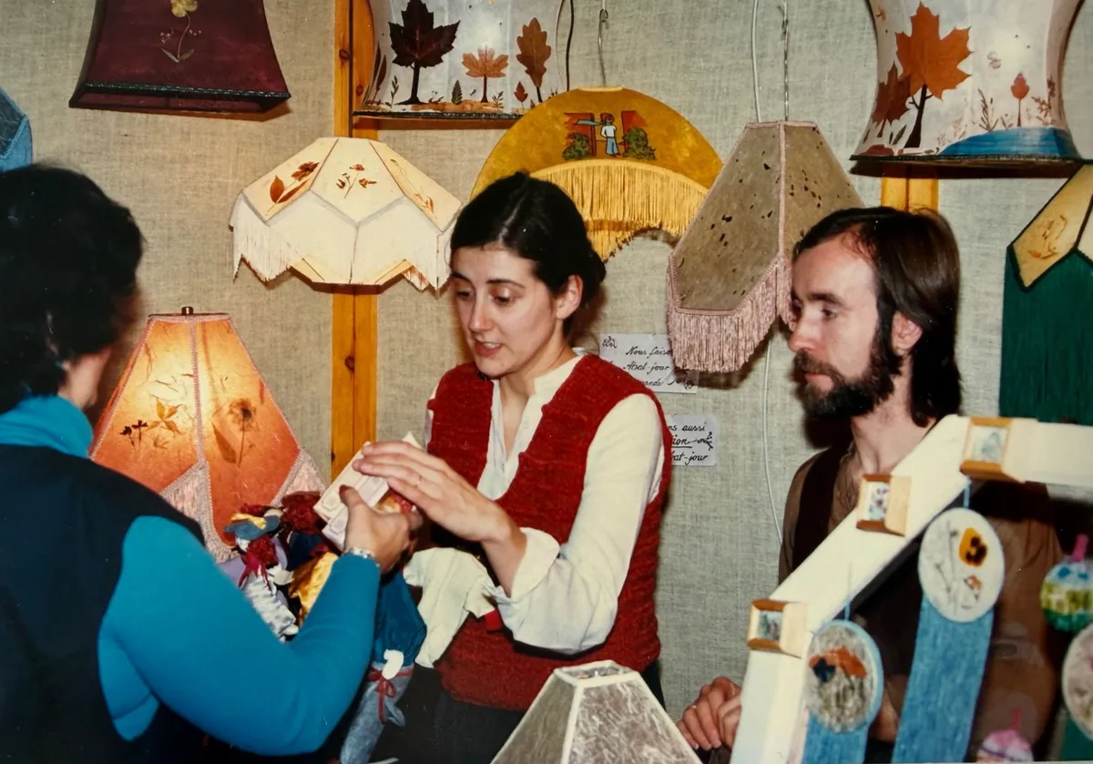 Grace et Manfred, jeunes, à leur kiosque aux Métiers d’Art de Lanaudière à Joliette au milieu des années 1980, montrant leur travail à une cliente, entourés de leurs abat-jour faits main décorés de feuilles et de fleurs pressées