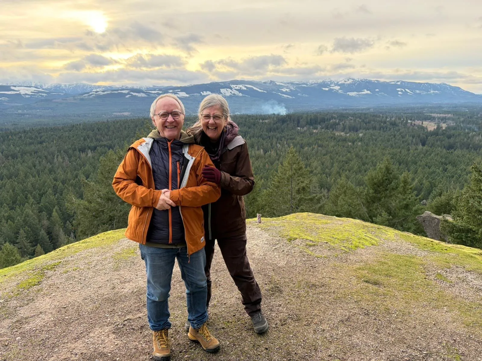 Manfred and Grace Hodder laughing on a mossy hilltop above a forested valley and snowy mountains